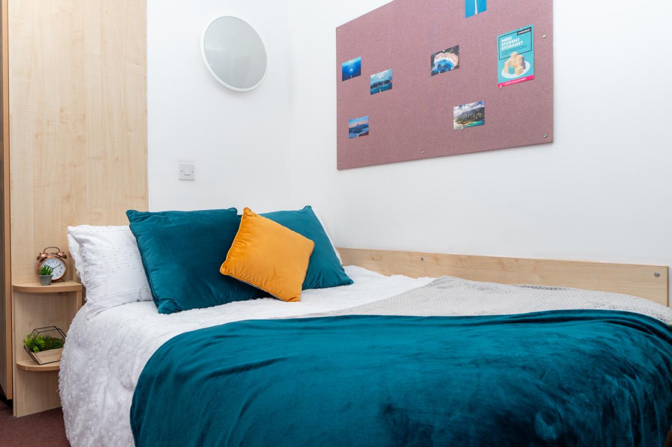 The corner of a bedroom showing a double bed with blue, yellow and white furnishings. There is a bedside table and a pinboard on the wall