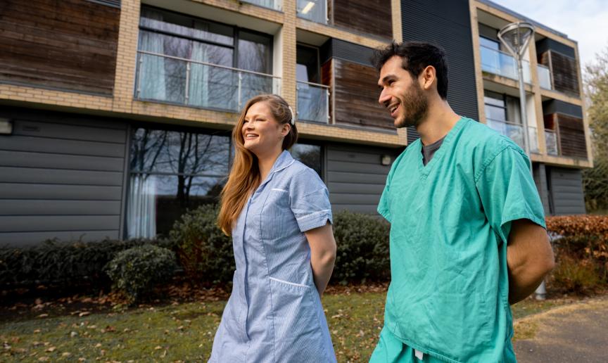 Two friends who live at Addenbrookes are walking outside the accommodation wearing their hospital work uniforms laughing and smiling together